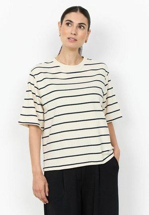Femme aux cheveux foncés en queue de cheval basse portant un haut crème à manches courtes avec des rayures horizontales noires et un pantalon noir, debout avec une main dans la poche.
