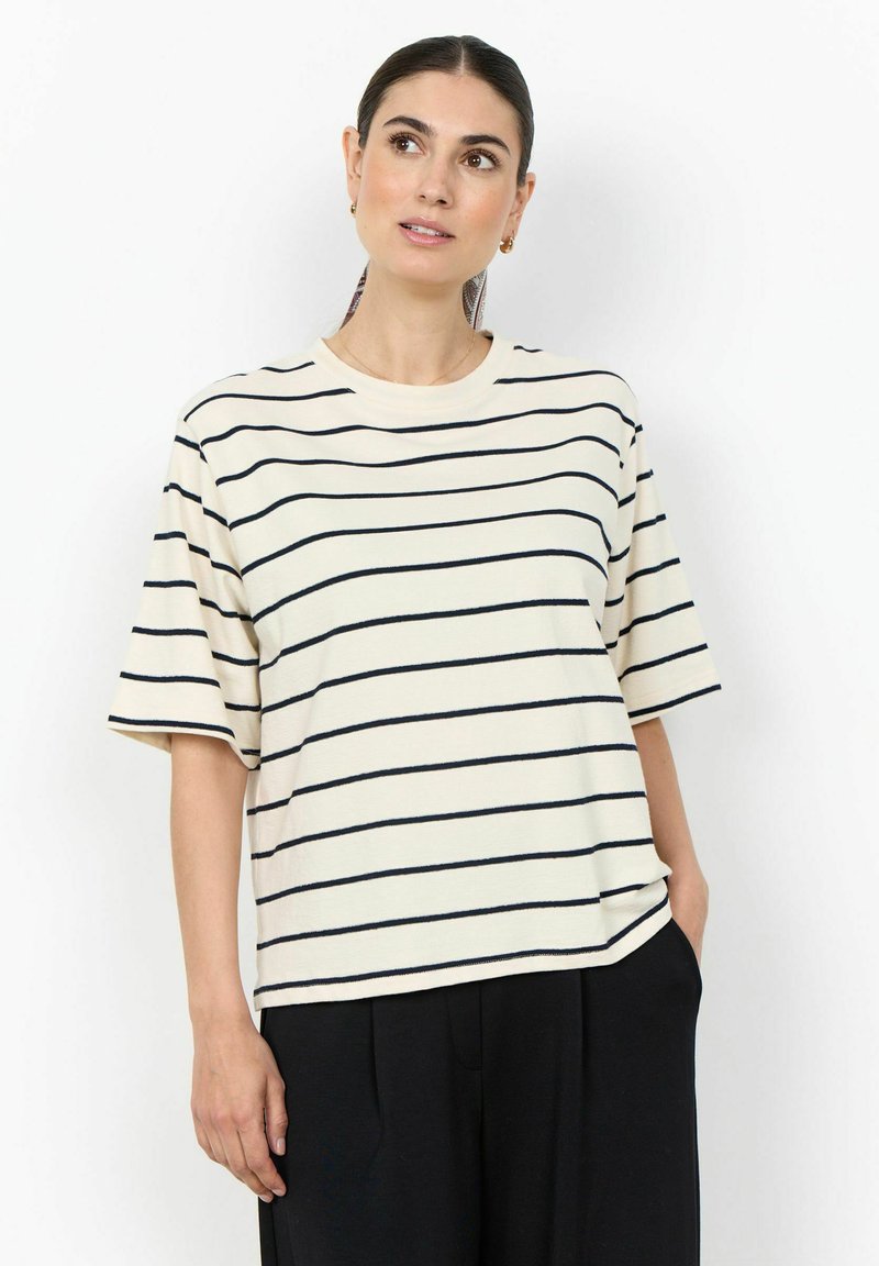 Femme aux cheveux foncés en queue de cheval basse portant un haut crème à manches courtes avec des rayures horizontales noires et un pantalon noir, debout avec une main dans la poche.