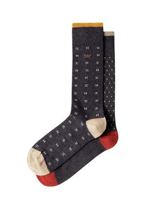 Dos calcetines gris oscuro con dedos beige, uno tiene pequeños patrones blancos en forma de H y un puño amarillo, el otro muestra un patrón de diamantes blanco y un talón rojo.