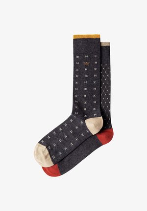 Dos calcetines gris oscuro con dedos beige, uno tiene pequeños patrones blancos en forma de H y un puño amarillo, el otro muestra un patrón de diamantes blanco y un talón rojo.