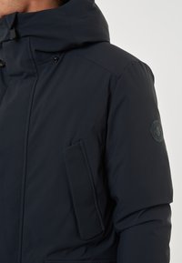 Giacca imbottita blu navy con cappuccio, dotata di tasca con zip sul petto, cuciture delle spalle arrotondate e una patch con logo sulla manica.
