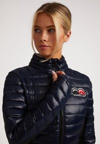Marineblaue Steppjacke mit hohem Kragen, dünnem horizontalem Steppmuster, Reißverschluss und einem bunten Logopatch auf der Brust.