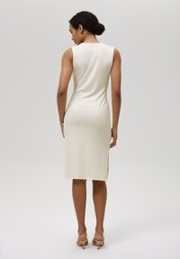 Knielang, ärmeloses Kleid aus leicht beigem, geripptem Stoff mit seitlichen Schlitzen. Glatte Textur und figurbetonter Silhouette. Minimalistisches Design.