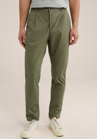 Pantalones ligeros de color verde oliva con frente plano, plisados y cierre de botón. Combinados con zapatillas de deporte blanco roto. Textura suave.