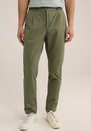 Pantalons légers, verts olive, avec un devant plat, des plis et une fermeture à bouton. Associés à des baskets blanc cassé. Texture lisse.