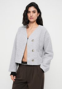 Γκρι κοντό σακάκι με υφή που μοιάζει με μάλλινο ύφασμα, διαθέτει τρία μεγάλα στρογγυλά κουμπιά και φαρδιά μανίκια. Χωρίς γιακά ή ορατό μοτίβο.