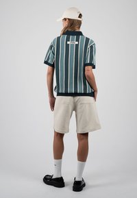 Persona que lleva una camiseta de manga corta a rayas verdes y blancas, pantalones cortos beige, calcetines blancos, zapatos negros y una gorra beige, de espaldas.