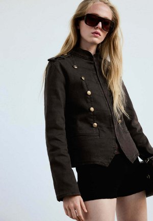 Joven mujer con largo cabello rubio, lleva gafas de sol grandes y oscuras y una chaqueta negra de estilo militar con botones dorados.
