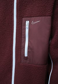 Burgunderfarget fleecejakke med en glatt nylonlomme, med en hvit glidelås og en liten Nike-logo i hvitt over lommen.