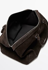 Tmavě hnědá semišová taška s velkým otevíráním, černou vnitřní podšívkou, stříbrným zipovým zapínáním a dvěma držadly pro nošení.