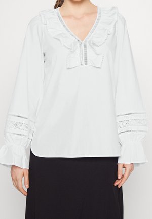 Vrouw draagt een witte blouse met lange mouwen, geplooide V-hals en kantdetail op de mouwen, gecombineerd met een zwarte rok.