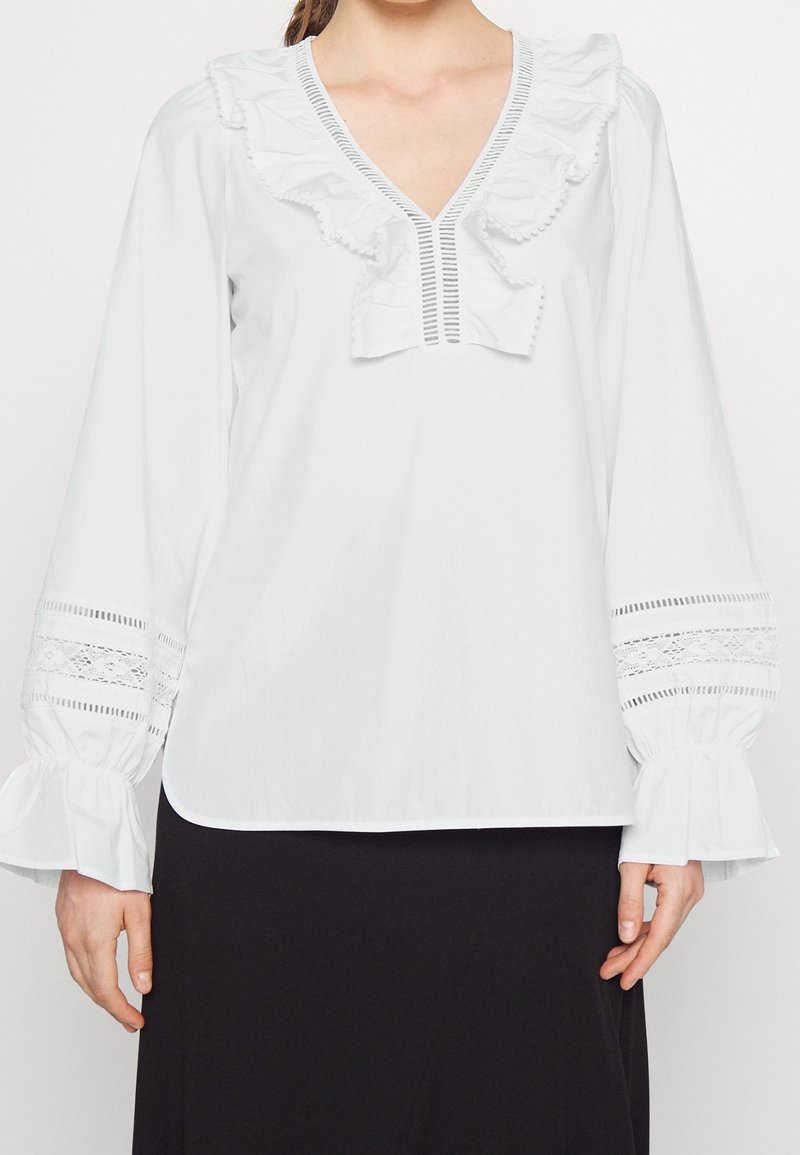Mujer lleva una blusa blanca de manga larga con escote en V con volantes y detalles de encaje en las mangas, combinada con una falda negra.