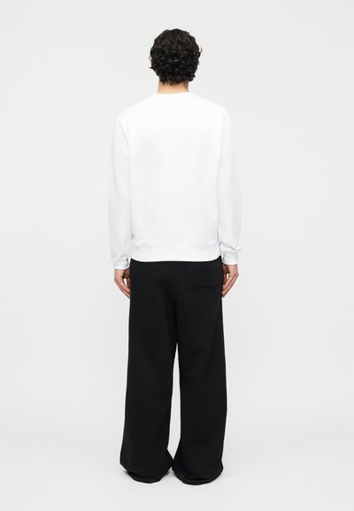 Άνδρας με σγουρά σκούρα μαλλιά στέκεται με την πλάτη, φορώντας λευκό μακρυμάνικο μπλουζάκι και άνετο μαύρο παντελόνι, πάνω σε απλό φόντο.