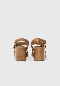 Bruna läderklackade sandaler med justerbara ankelremmar, en bred blockklack och korsremdesign, med detaljer i guldfärgad metall.