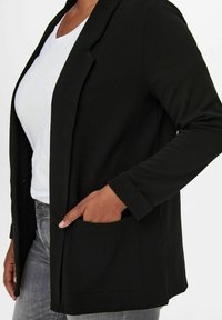 Musta bleiseri, jossa on teksturoitu kangas, avoin edusta, pitkät hihat ja yksi sivutasku. Päällä valkoinen toppi ja harmaat farkut.