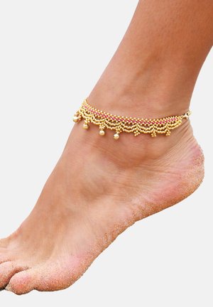 Goldener Fußkettchen mit Perlendekor und rosa Akzenten sowie kleinen hängenden Glöckchen, getragen an einem nackten Fuß, der teilweise mit Sand bedeckt ist.