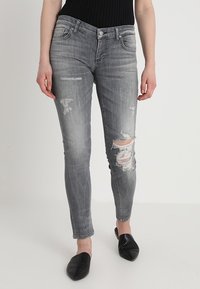 Jeans skinny de denim gris con parches desgastados y roturas en las rodillas. Presentan un diseño clásico de cinco bolsillos y una cremallera en la bragueta.