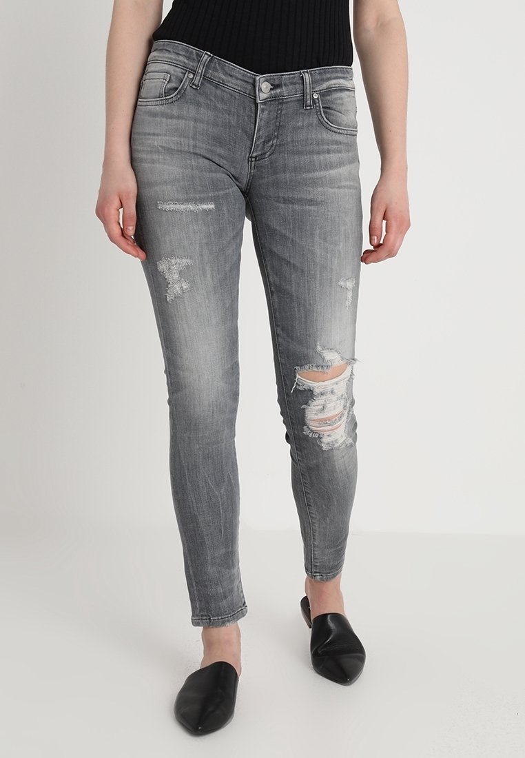 Jeans skinny de denim gris con parches desgastados y roturas en las rodillas. Presentan un diseño clásico de cinco bolsillos y una cremallera en la bragueta.