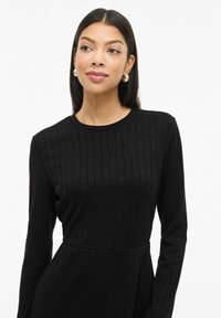 Robe noire ajustée à manches longues, avec une texture côtelée et un col rond. Elle présente un détail à la taille et un tissu lisse pour le confort.