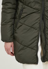 Ruka spočívá na boku, oblečená v tmavě zelené prošívané bundě se dvěma zipovými kapsami a částečně rozepnutým předním zipem přes bílé kalhoty.