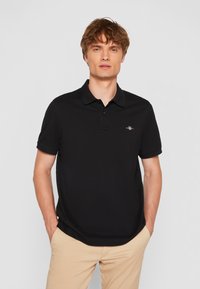 Must na kortsete varrukatega polo särk, millel on nööpidega krae ja rindkere kohal väike broderiangiga logo. Materjal näeb välja sile ja kerge.