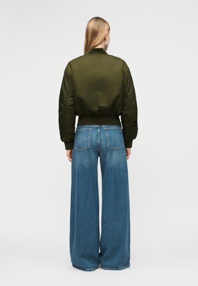 Γυναίκα με μακριά ξανθά μαλλιά, φορώντας πράσινο μπουφάν τύπου bomber και ψηλόμεσα ευρύχωρα μπλε τζιν παντελόνια, στέκεται με την πλάτη προς το μέρος μας.