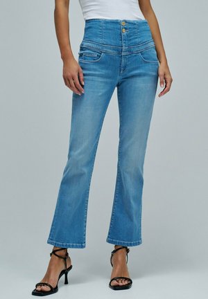 Femme portant un jean évasé taille haute bleu clair avec fermeture à double bouton, associé à des sandales à talons noires à brides et un haut blanc.