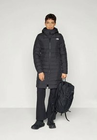 Black puffer coat with horizontal stitching, zip closure, and hood, worn with black pants and sneakers, alongside a black backpack.