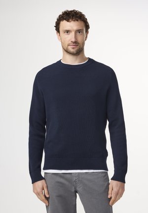 Pull en tricot côtelé navy à manches longues et col rond, superposé à une chemise blanche, associé à un pantalon gris clair. Design simple.