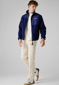 Marine fleecejack met contrasterende blauwe nylon bovenkant, ritszak en rode rits. Gecombineerd met beige cargobroek en witte sneakers.