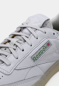 White leather sneaker with grey laces, featuring a green "Reebok" logo and British flag. Rounded shape with padded tongue and thin sole.