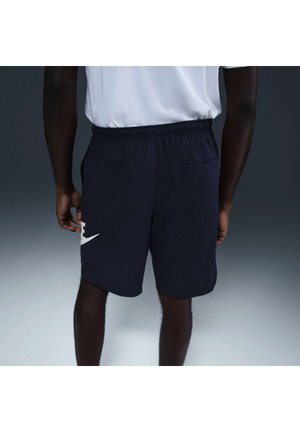 Person iført sorte træningsshorts og hvidt skjorte set bagfra mod en ensfarvet grå baggrund.