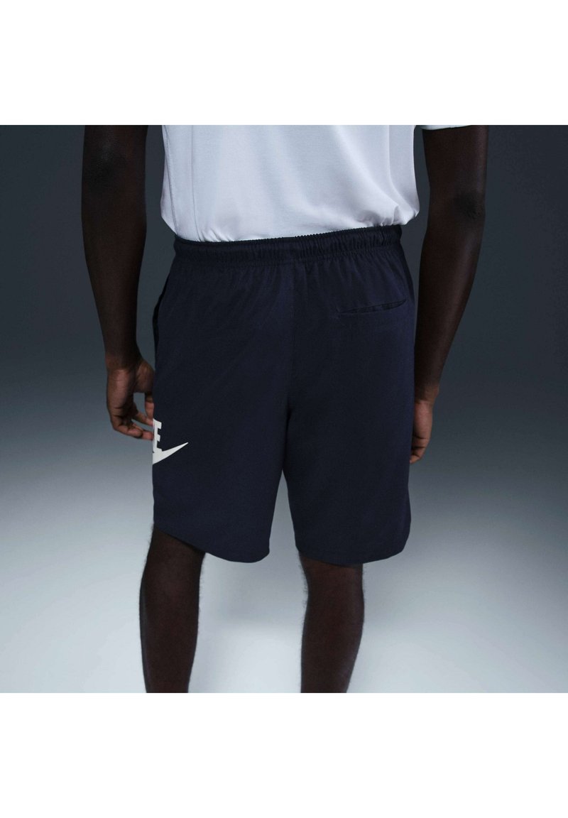 Persona vestida con pantalones cortos deportivos negros y camiseta blanca vista desde atrás contra un fondo gris liso.