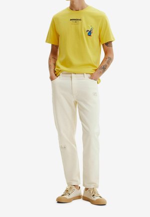 Gelbes Baumwoll-T-Shirt mit "Desigual"-Logo und buntem Pflanzenmotiv, kombiniert mit cremefarbener Jeans und beigefarbenen Sneakers.