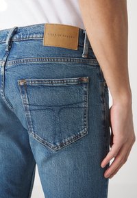 Modré džínové kalhoty s zadní kapsou, kožený štítek s nápisem "TIGER OF SWEDEN", kontrastní švy a mírně opotřebovaný vzhled.