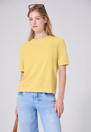 Jeune femme avec des lunettes de soleil sur la tête, portant un haut jaune à manches courtes, un jean bleu clair, des créoles et tenant un sac à main marron.