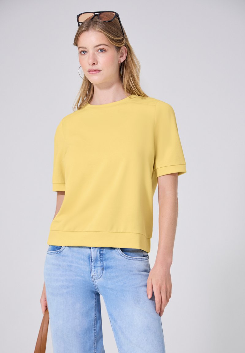 Jeune femme avec des lunettes de soleil sur la tête, portant un haut jaune à manches courtes, un jean bleu clair, des créoles et tenant un sac à main marron.