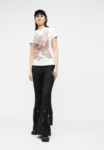 Νεαρή γυναίκα με λευκό μπλουζάκι με σχέδιο, μαύρο cargo παντελόνι με λουριά και φερμουάρ, μαύρα παπούτσια και μαύρο καπέλο μπέιζμπολ, όρθια.