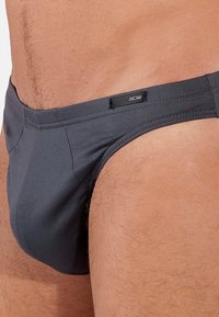 Dunkelgraue Schwimmbriefs mit glatter Struktur, die ein enges Design und ein kleines Logo-Etikett am Taillenbund aufweisen.