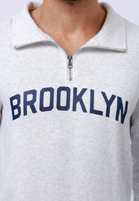 Světle šedá mikina na zip s límečkem, která má na hrudi velký modrý grafický nápis "BROOKLYN". Měkký materiál s viditelnou texturou.