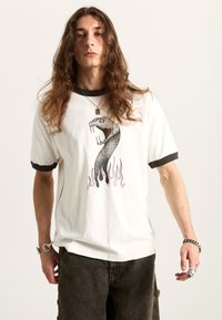 Camiseta blanca de algodón con cuello negro, que presenta un gráfico de serpiente y un diseño de llamas rosas. Modelo combinado con pantalones oscuros.