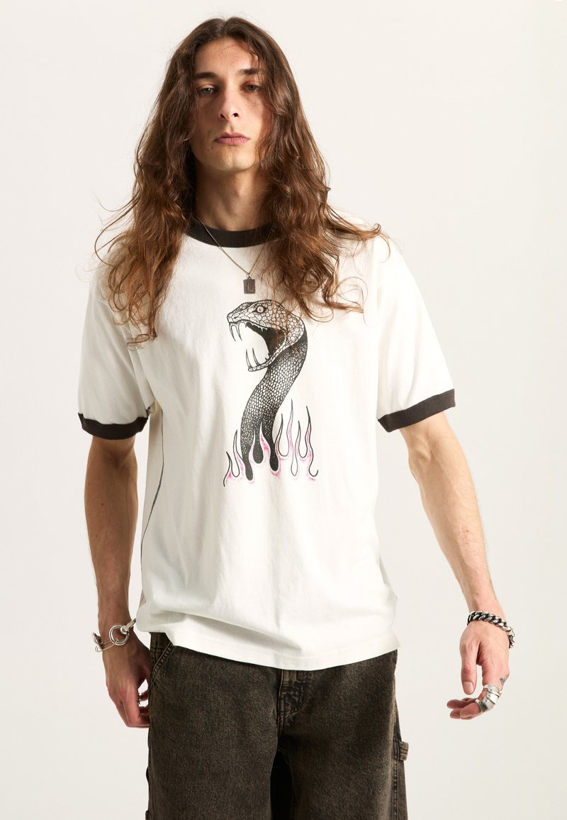 Camiseta blanca de algodón con cuello negro, que presenta un gráfico de serpiente y un diseño de llamas rosas. Modelo combinado con pantalones oscuros.