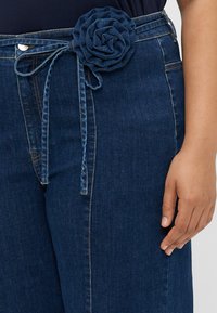 Calças de ganga com um tom azul escuro, apresentando uma cintura atada decorada com um detalhe de rosa em tecido e costuras contrastantes.