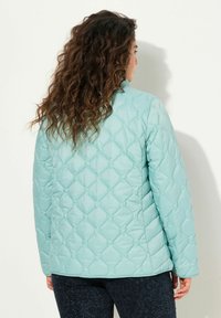 Giacca trapuntata blu chiaro con una texture liscia, dotata di colletto alto e motivo a diamante, indossata con pantaloni scuri a fantasia.