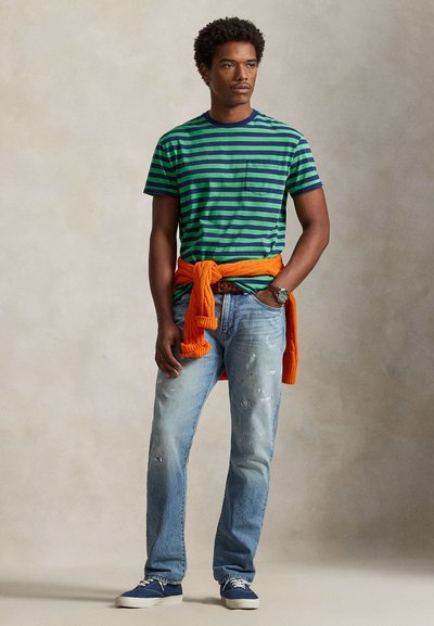 T-shirt à manches courtes rayé vert et marine avec une poche poitrine, associé à un jean bleu délavé. Un pull orange est noué autour de la taille.