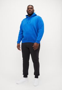 Casaco capuz azul com bolso frontal e cordão, combinado com calças de fato pretas. Modelo usa sapatilhas brancas. Textura suave, corte relaxado.