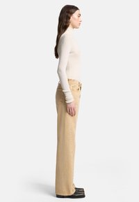 Beżowe jeansy o szerokich nogawkach z wysokim stanem, zestawione z kremowym prążkowanym golfem. Modelka widziana z boku, ukazująca dopasowaną sylwetkę.
