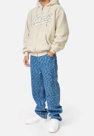 Beiger Fleece-Kapuzenpullover mit Reißverschluss und vorderem Logo, kombiniert mit blauen, gemusterten, locker sitzenden Jeans und weißen Sneakers. Lässiger Streetwear-Stil.