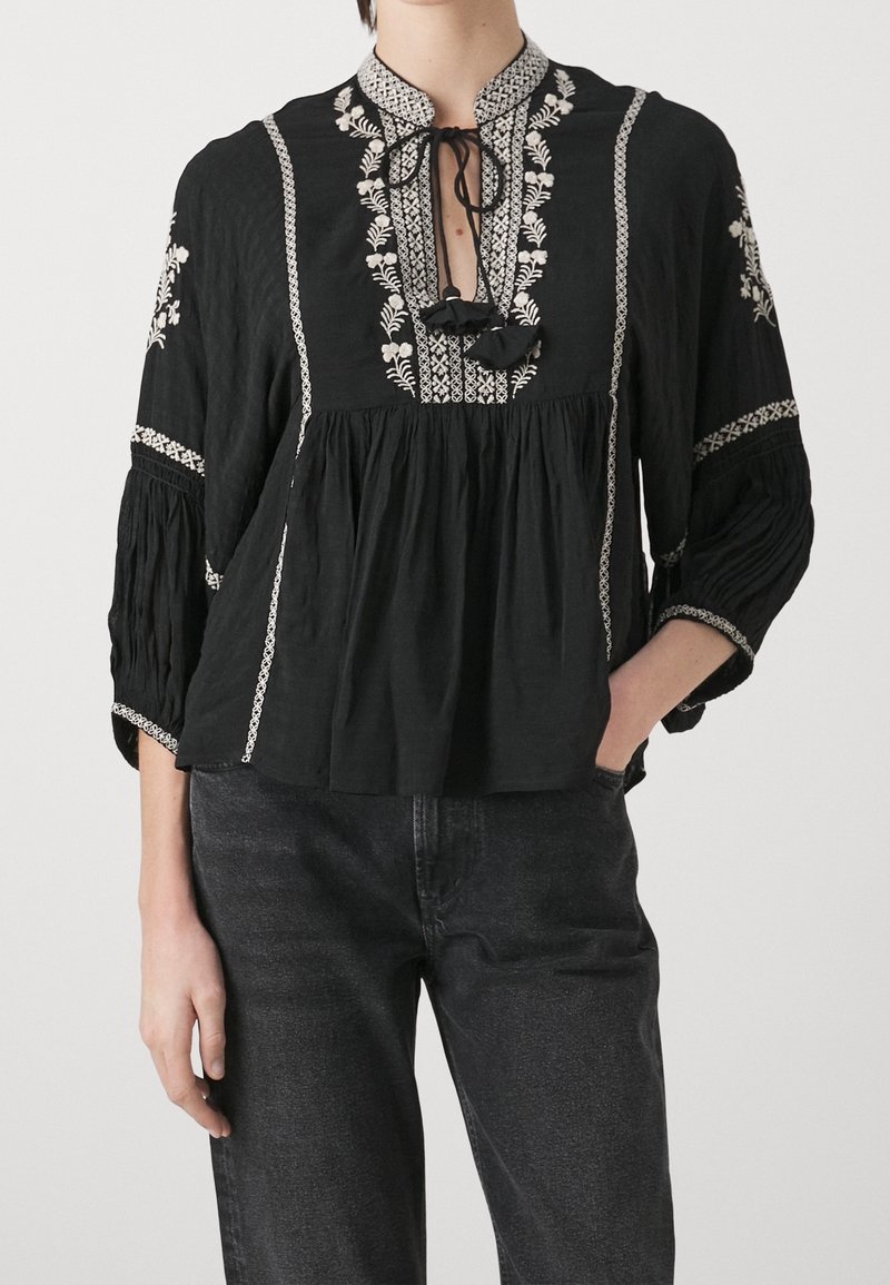 Blouse noire avec broderie florale blanche sur le devant, les manches et le col, dotée d'un col à nouer et de manches amples 3/4.