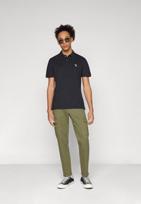 Polo negro de manga corta con un pequeño logo, combinado con pantalones cargo verde oliva que cuentan con múltiples bolsillos y zapatillas negras.